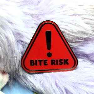 May include: A red enamel pin in the shape of a warning triangle with a black border. The pin features a black exclamation point and the words "BITE RISK" in white. The pin is set against a backdrop of fluffy, light purple faux fur.