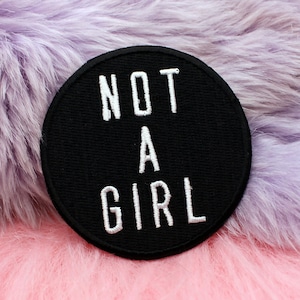 Pode incluir: Um remendo redondo preto com texto branco que diz "NOT A GIRL" em um fundo difuso roxo e rosa.