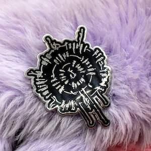 May include: A black and silver pin with an abstract design, set against a backdrop of fluffy, lavender-coloured faux fur. The pin's design features a central spiral surrounded by radiating lines and shapes.