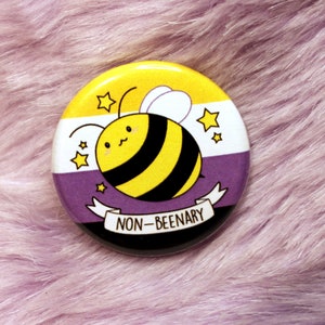 May include: A yellow, black, and white button with a cartoon bee and the text "NON-BEENARY" on a purple and white fuzzy surface.