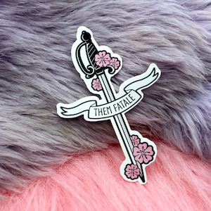 May include: A black sticker with a sword and flowers. The sword has a white ribbon with the text "Them Fatale" on it.