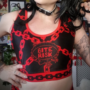 May include: Black and red crop top with a chain design and the words "BITE RISK". The top has a red trim and is worn with a black collar. The person in the image has black hair and is wearing a nose ring.