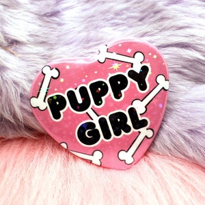 Peut inclure: Épinglette en forme de cœur rose avec des motifs d'os blancs et le texte "Puppy Girl" en lettres noires.
