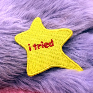 Può includere: Una toppa gialla a forma di stella con la scritta ricamata "i tried" in rosso. La toppa è posta su uno sfondo di soffice finta pelliccia nei toni del viola e del rosa.