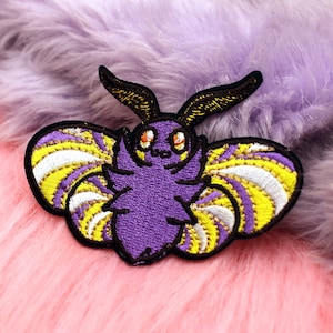 May include: Embroidered patch featuring a stylized moth design. The moth has a purple body, yellow and white striped wings, and black antennae. The patch is outlined in black and set against a pink and purple fluffy background.