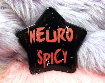 Può includere: Uno spillo nero a forma di stella con le parole "Neuro Spicy" in lettere rosse. Lo spillo è su una superficie pelosa viola e rosa.