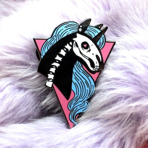 Puede incluir: Un pin de esmalte negro y blanco de un unicornio esqueleto con pelo azul, sobre un fondo de triángulo rosa.