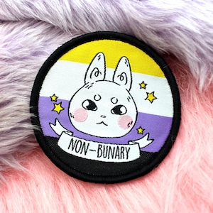 May include: A round patch with a black border featuring a cartoon bunny with pink cheeks and a banner that reads "NON-BINARY". The background has yellow, white, and purple stripes with gold stars.