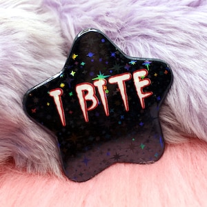 Könnte beinhalten: Eine schwarze sternförmige Anstecknadel mit glitzernder Oberfläche und dem Text "I Bite" in Regenbogenfarben.