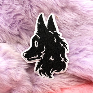 May include: A black and white embroidered patch depicting a wolf's head. The patch has a white border and is set against a backdrop of fluffy, lavender and pink faux fur.