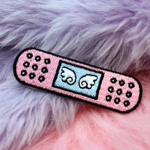 May include: Pink fabric patch shaped like a bandage with white wings and black stitching. The patch is on a pink and purple fuzzy surface.