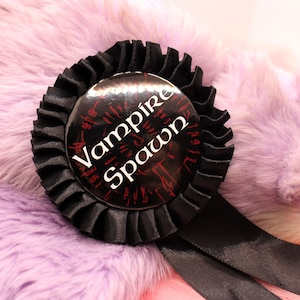 Könnte beinhalten: Schwarz-weißer Knopf mit dem Text "Vampire Spawn" auf einem violetten, flauschigen Hintergrund.
