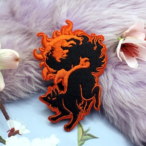 May include: An embroidered patch featuring a black wolf silhouette positioned below a stylized orange sun with a face. The design uses vibrant orange thread for the sun's rays and the wolf's outline, contrasting against the solid black thread fill.