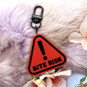 Op de afbeelding: Een heldere acryl sleutelhanger met een zwarte karabijnhaak. De sleutelhanger heeft een rode waarschuwingsdriehoek met een zwart uitroepteken en de woorden "BITE RISK" in witte letters. De achtergrond is een pluizig, lavendelkleurig materiaal.