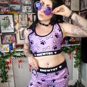 May include: A lilac sports bra and shorts set with a black waistband. The set features a pattern of black paw prints and white bones. The waistband of both pieces reads "CHEWTOY" in white lettering.