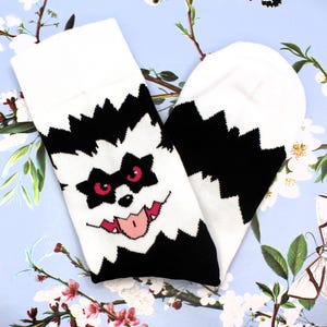 May include: White socks with a black and white monster face design. The monster has red eyes and a pink tongue. The socks are on a light blue floral background.