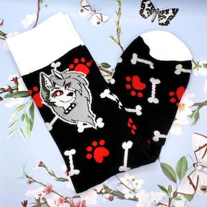 May include: Black socks with a white cuff featuring a cartoon wolf head, red paw prints, and white bones. The wolf has gray fur and red eyes. The socks are on a light blue floral background.