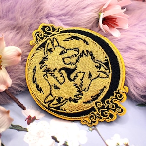 May include: A circular embroidered patch featuring a golden design of three wolf heads arranged in a triangular formation against a black background, with decorative swirling border elements at the top and bottom edges of the circular frame.