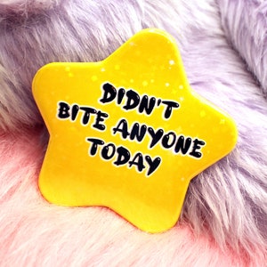 Könnte beinhalten: Ein gelber sternförmiger Pin mit dem Text "Didn't Bite Anyone Today" in schwarzer Schrift. Der Pin hat eine glitzernde Oberfläche.