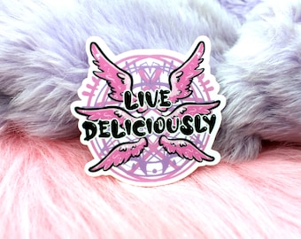 Può includere: Un adesivo rosa e bianco con un design circolare e due ali rosa. Il testo "LIVE DELICIOUSLY" è scritto in lettere nere.