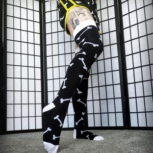 May include: Black thigh-high socks with a white bone pattern. The socks are paired with black shorts with yellow trim and the word "NERVOUS" on the waistband. The socks have white toes and heels.