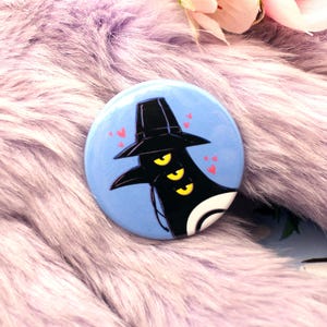 May include: A round, light blue button with a cartoon illustration of a black figure wearing a hat and three yellow eyes. Small pink hearts are scattered around the figure. The button is set against a fluffy, purple background.