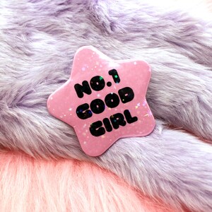 Puede incluir: Bot&oacute;n rosa en forma de estrella con el texto "No. 1 Good Girl" en letras negras. El bot&oacute;n est&aacute; sobre una superficie peluda de color morado claro.