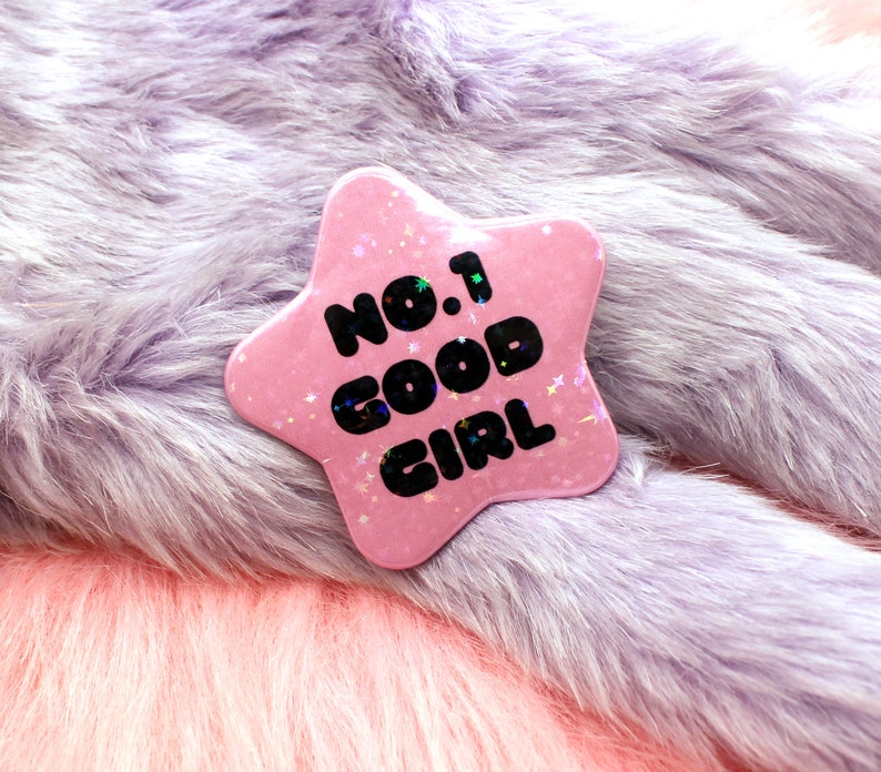 Puede incluir: Bot&oacute;n rosa en forma de estrella con el texto "No. 1 Good Girl" en letras negras. El bot&oacute;n est&aacute; sobre una superficie peluda de color morado claro.