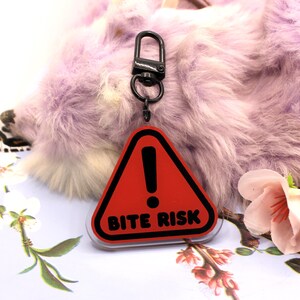 This 5cm triangluar acrylic charm is the perfect addition to any keyring or ita bag! The design features a triangluar red warning sign with the text '! BITE RISK' in large letters. It is double sided with a lobster clasp for easy fastening.