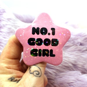 Puede incluir: Bot&oacute;n rosa en forma de estrella con purpurina y el texto "No.1 Good Girl" en negro.