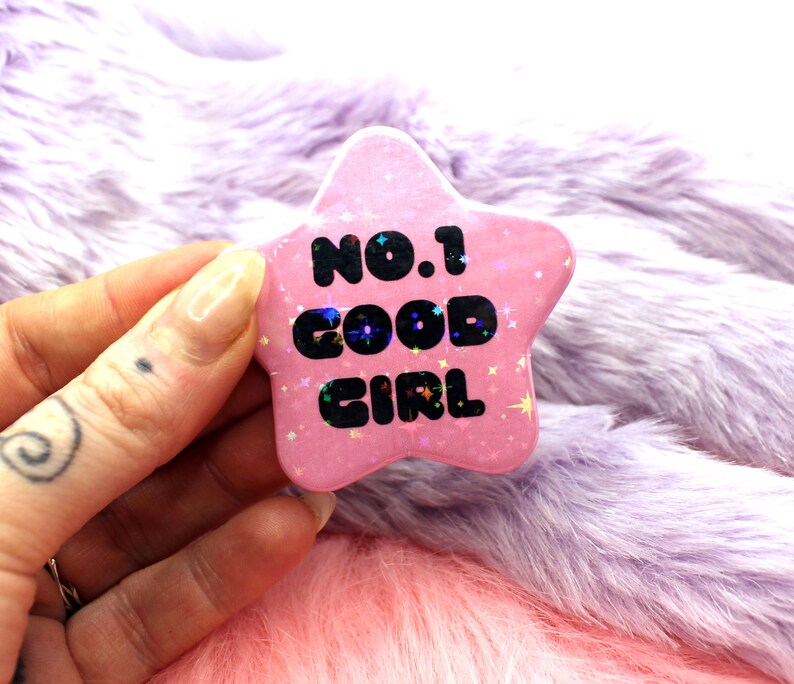Puede incluir: Pin de estrella rosa con purpurina y el texto "NO.1 GOOD GIRL" en negro.