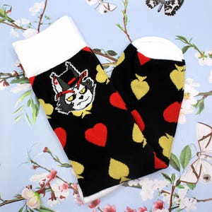 May include: A pair of black socks with a white cuff and toe. The socks feature a cartoon cat wearing a top hat and playing card symbols in red and gold. The socks are on a light blue floral background.