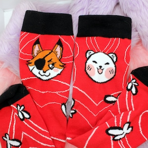 Puede incluir: Un par de calcetines rojos con líneas blancas y un puño negro. Un calcetín presenta un zorro de dibujos animados con un parche en el ojo y el otro presenta un oso de dibujos animados con mejillas sonrosadas.