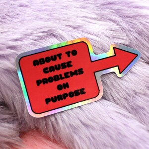 May include: A red and silver holographic sticker with an arrow pointing to the right. The sticker says "About to cause problems on purpose."