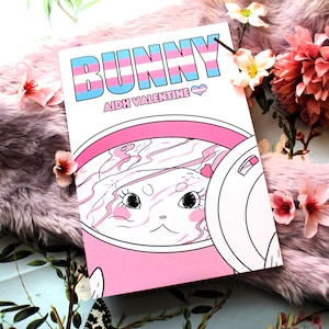 Puede incluir: Un cómic rosa y blanco con el título "BUNNY" en colores arcoíris y el texto "AIDH VALENTINE" debajo. La portada presenta un conejito de dibujos animados en una taza rosa con un patrón de remolino rosa y blanco.