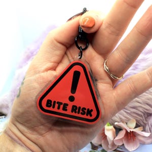 This 5cm triangluar acrylic charm is the perfect addition to any keyring or ita bag! The design features a triangluar red warning sign with the text '! BITE RISK' in large letters. It is double sided with a lobster clasp for easy fastening.