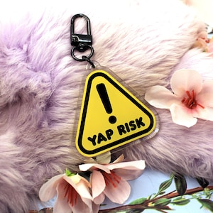 May include: A yellow and black keychain with the text "YAP RISK" inside a warning triangle. The keychain has a black clasp and is set against a fluffy, lavender background with pink flowers.