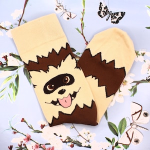 May include: A pair of novelty socks with a cartoon monster face design. The socks are primarily beige with brown accents and a pink tongue. The monster face is centered on the front of the socks, with a jagged brown pattern around the face.