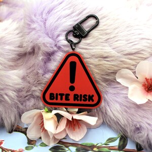 This 5cm triangluar acrylic charm is the perfect addition to any keyring or ita bag! The design features a triangluar red warning sign with the text '! BITE RISK' in large letters. It is double sided with a lobster clasp for easy fastening.