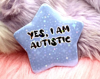 Può includere: Un bottone blu a forma di stella con sfondo glitterato e il testo "YES, I AM AUTISTIC" in nero.