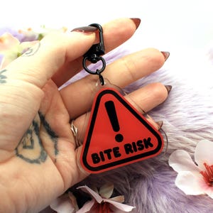 This 5cm triangluar acrylic charm is the perfect addition to any keyring or ita bag! The design features a triangluar red warning sign with the text '! BITE RISK' in large letters. It is double sided with a lobster clasp for easy fastening.