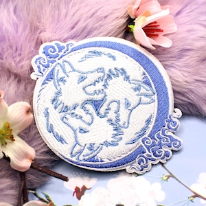May include: A circular embroidered patch featuring a white and blue design of three wolf heads arranged in a circle. The patch has a blue border and decorative cloud-like patterns on the edges, rendered in shades of white and blue thread.