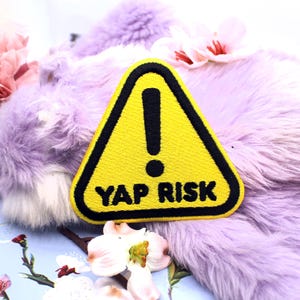 Könnte beinhalten: Ein gelber Aufnäher mit schwarzem Rand und Ausrufezeichen, mit den Worten "YAP RISK" in Schwarz. Der Aufnäher ist dreieckig und liegt auf lila Kunstfell, mit rosa und weißen Blumen.