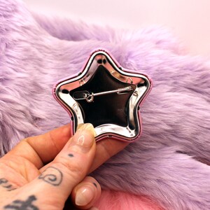 Puede incluir: Un pin de estrella plateado con un borde rosa. El pin tiene un cierre de seguridad en la parte posterior.