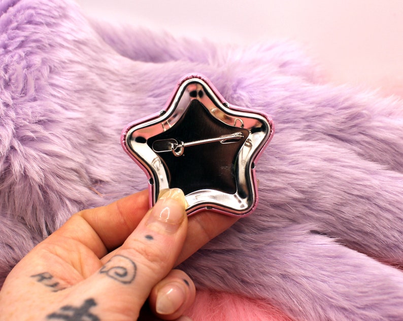 Puede incluir: Un pin de estrella plateado con un borde rosa. El pin tiene un cierre de seguridad en la parte posterior.