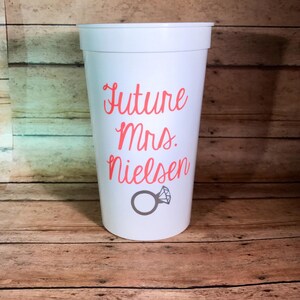 May include: White plastic cup with the text "Future Mrs. Nielsen" in pink lettering and a gray ring with a diamond on top.