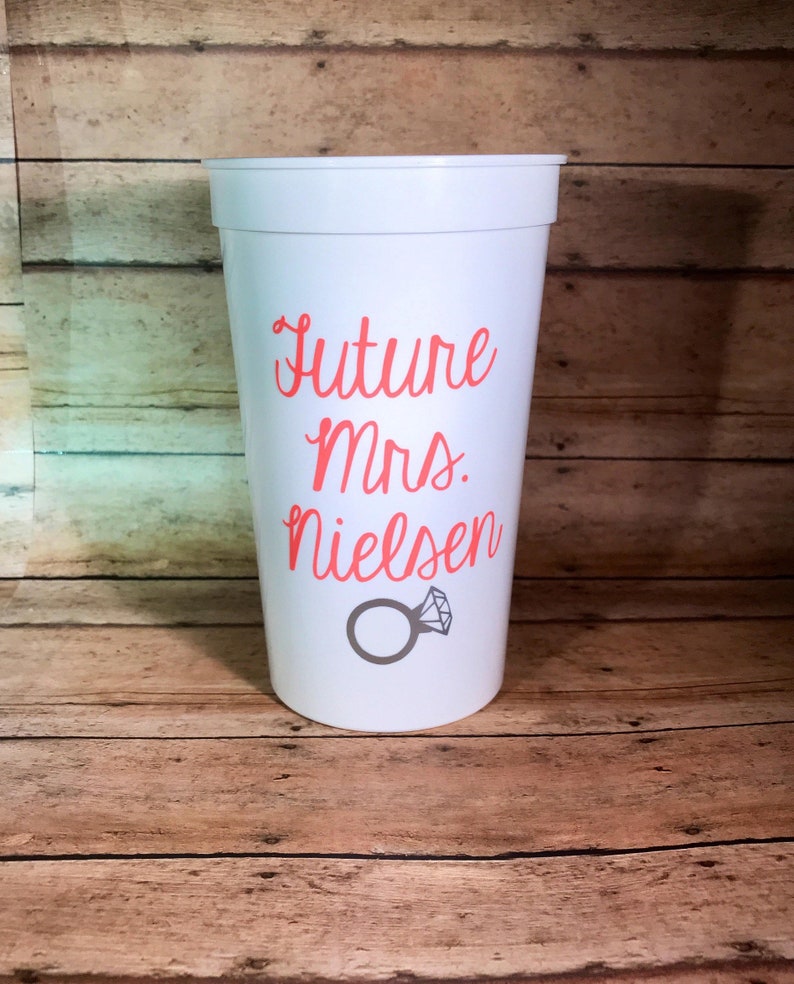 May include: White plastic cup with the text "Future Mrs. Nielsen" in pink lettering and a gray ring with a diamond on top.