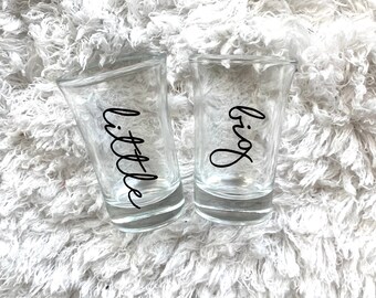 May include: Two clear glass shot glasses with black lettering. One glass says "big" and the other says "little".