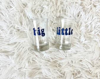 May include: Two clear glass shot glasses with blue lettering. One glass says "big" and the other says "little".