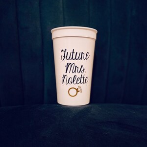 May include: White plastic cup with the text "Future Mrs. Nolette" and a gold ring with a diamond on it.